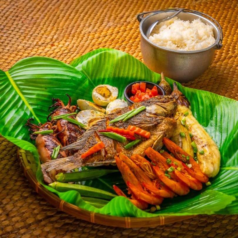 Filipino grilled seafood platter with fish, squid, shrimp, vegetables, and rice on banana leaves.