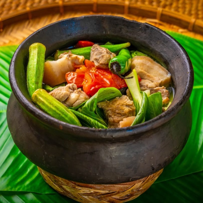 Filipino Sinigang in a black pot with meat, tomatoes, okra, and green peppers on a banana leaf.