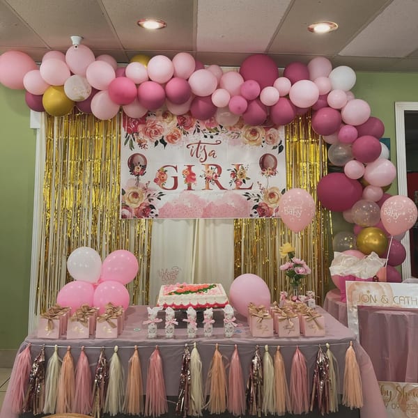 A baby shower setup with pink and gold balloons, a cake, and "It's a Girl" banner at Nipa Hut.