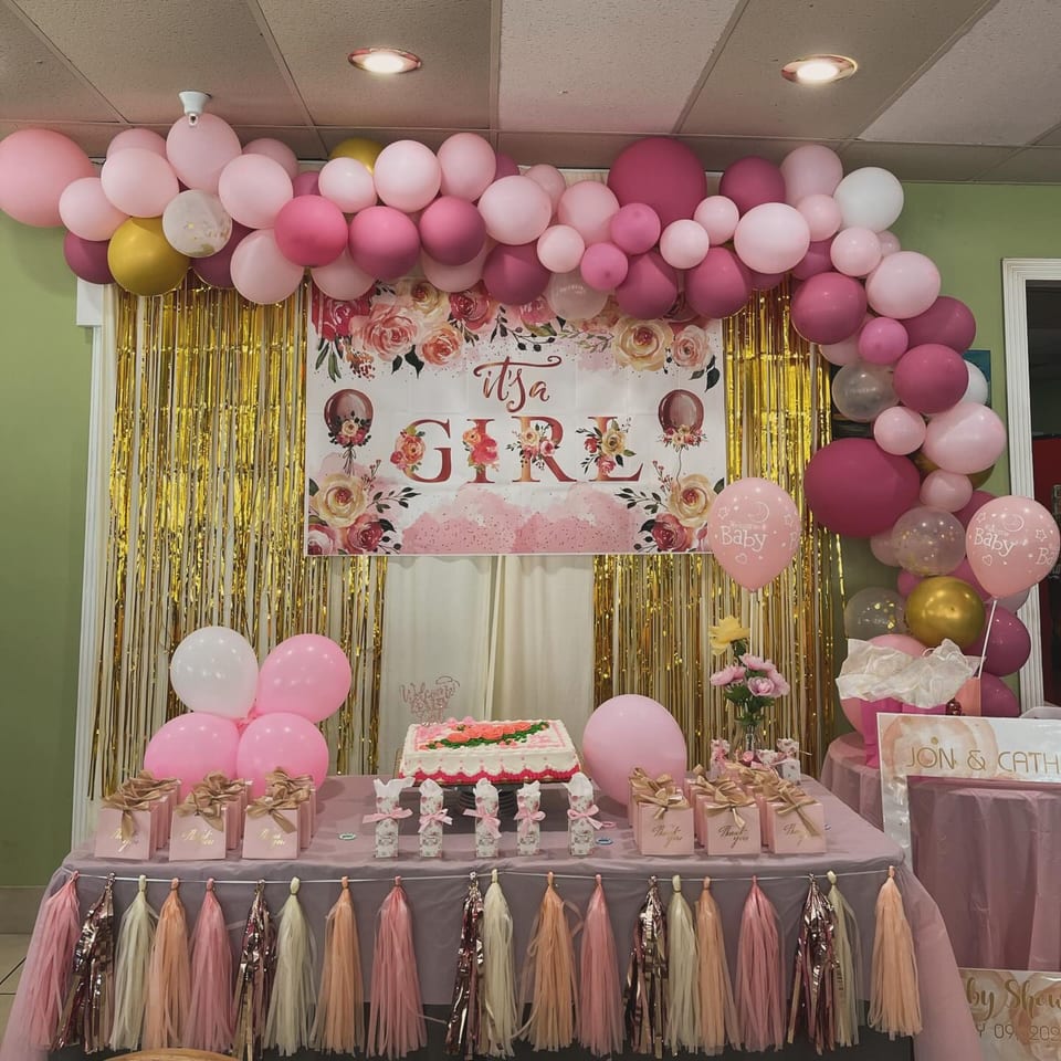 A baby shower setup with pink and gold balloons, a cake, and "It's a Girl" banner at Nipa Hut.