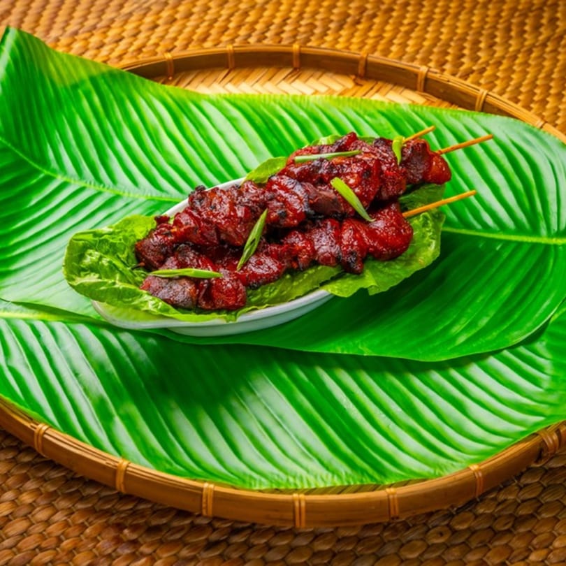 Filipino pork skewers, or 'Pork BBQ,' on a bed of lettuce and banana leaves, served on a woven tray.